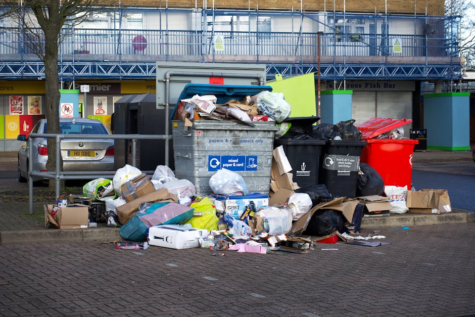 A large pile of mixed waste, including crumpled cardboard boxes, plastic bags, paper, and discarded packaging, is overflowing from a collection bin situated on a paved parking area. The waste items va