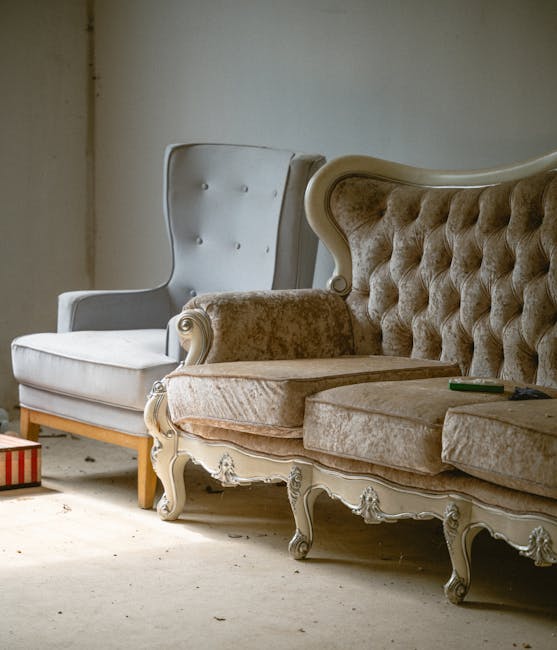 A person standing on a wooden floor is lifting the corner of a grey upholstered sofa, revealing the wooden leg structure beneath. The individual is dressed in dark pants and a grey sweater, with their