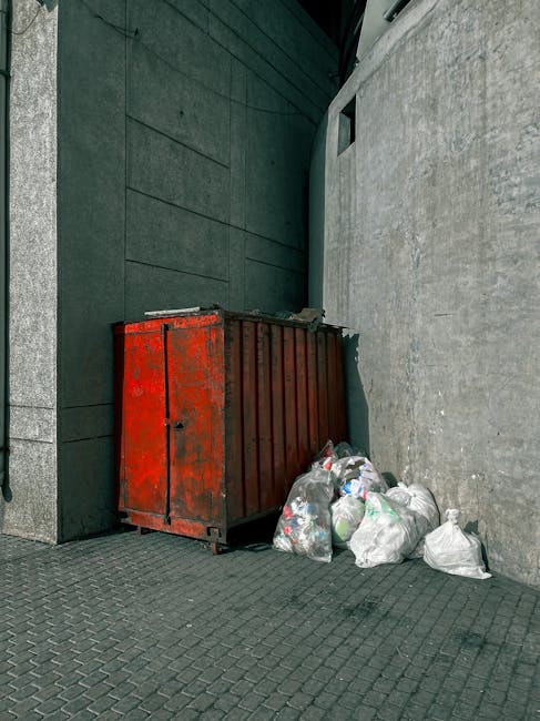 A person wearing light blue jeans and white sneakers is crouching on a paved outdoor surface, holding a collection of small waste items in their hand. The waste includes a crumpled white paper cup, so